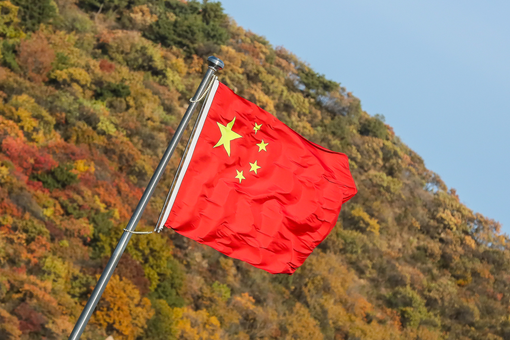 Bandeira da China hasteada e tremulando ao vendo na Grande Muralha, próximo a Pequim; ao fundo montanha coberta por vegetação e céu azul claro (Foto: Isac Nóbrega/PR)