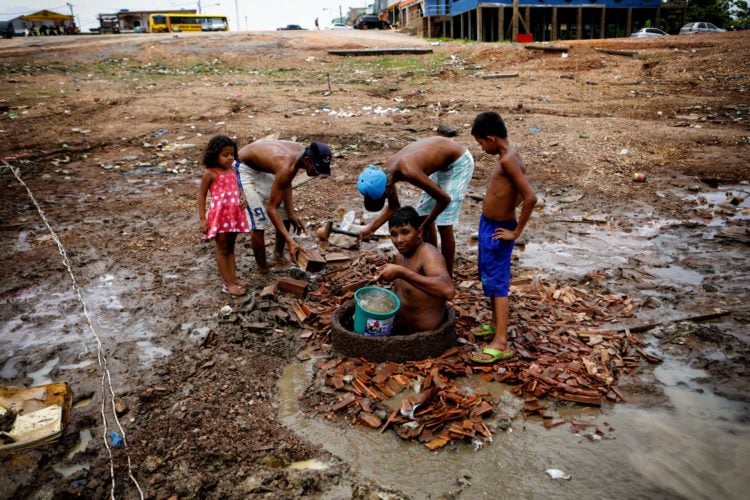 Diários da COP26: Clima tem que incorporar dimensões de classe, etnia e gênero. Na imagem: Homem adulto retira balde de água de uma cisterna em chão de terra batida, com quatro crianças ao redor, todos em situação de pobreza extrema. Cenário gerado por secas, queimadas e enchentes atípicas, que deixam populações tradicionais da Amazônia em situação de alta vulnerabilidade social e econômica (Foto: Joel Rosa/Amazônia Real)