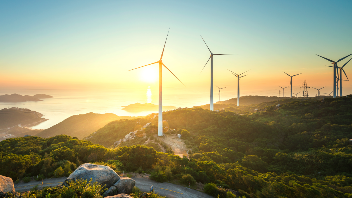 Inflação eleva margens para renováveis em 2022, indica relatório da Platts. Na imagem: Turbinas eólicas sobre montanhas cobertas com vegetação e, ao fundo, sol brilhando se reflete no oceano (Foto: Reprodução Twitter EY)