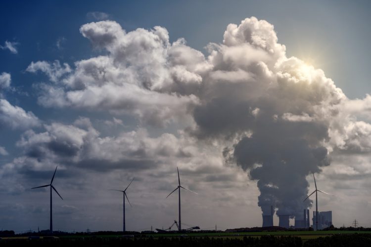 Obrigações das distribuidoras de combustíveis e CBIOs: desafios da regulamentação do mercado de carbono. Na imagem: Planta termelétrica emitindo grande volume de emissões, próximo a aerogeradores, ao fundo (Foto: Joe/jplenio/Pixabay)