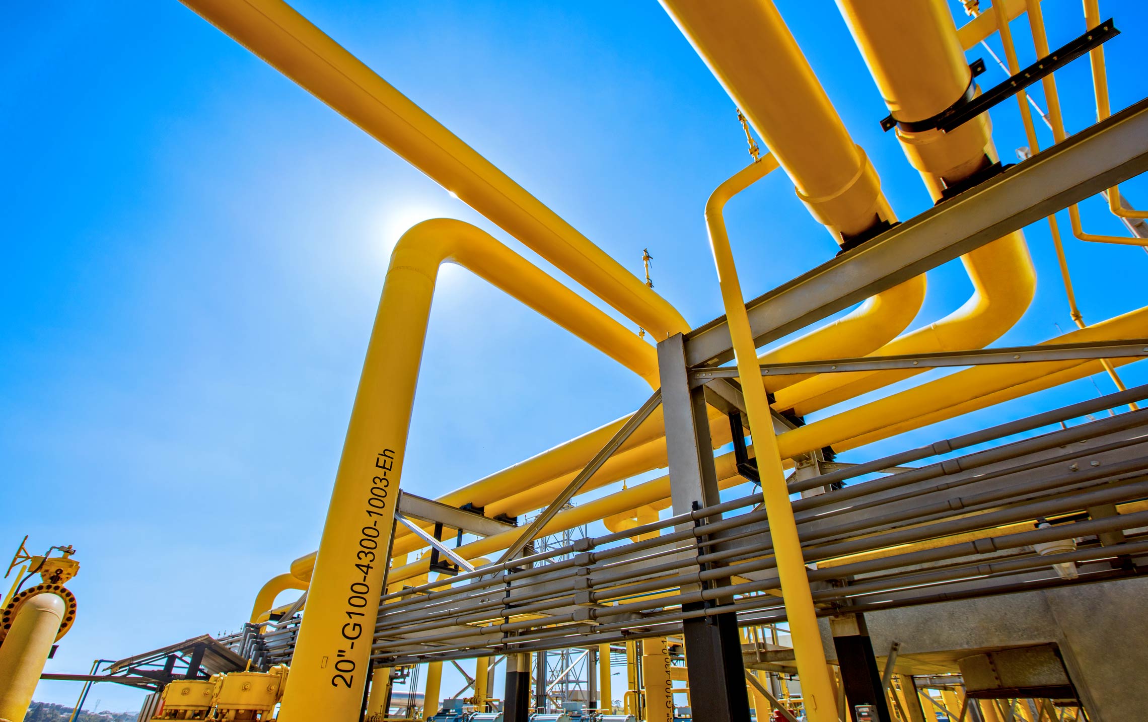 A importância da integração do gás no Brasil. Na imagem: Visão de baixo pra cima de instalações em gasoduto da NTS, com tubulações metálicas em amarelo, e céu azul ao fundo (Foto: Divulgação)