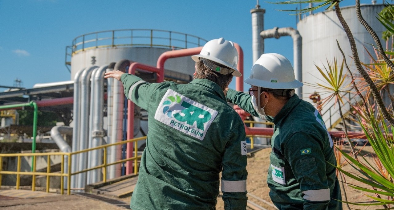 O que o contrato de processamento de gás com a Petrobras significa para a 3R Petroleum. Na imagem: Dois trabalhadores em planta da 3R Petroleum. Vestem macacão verde escuro com sinalizações de segurança e logo escrito "3R Petroleum", com destaque, nas costas. Usam capacete branco. Um deles aponta para as instalações com o braço esquerdo estendido (Foto: Divulgação)