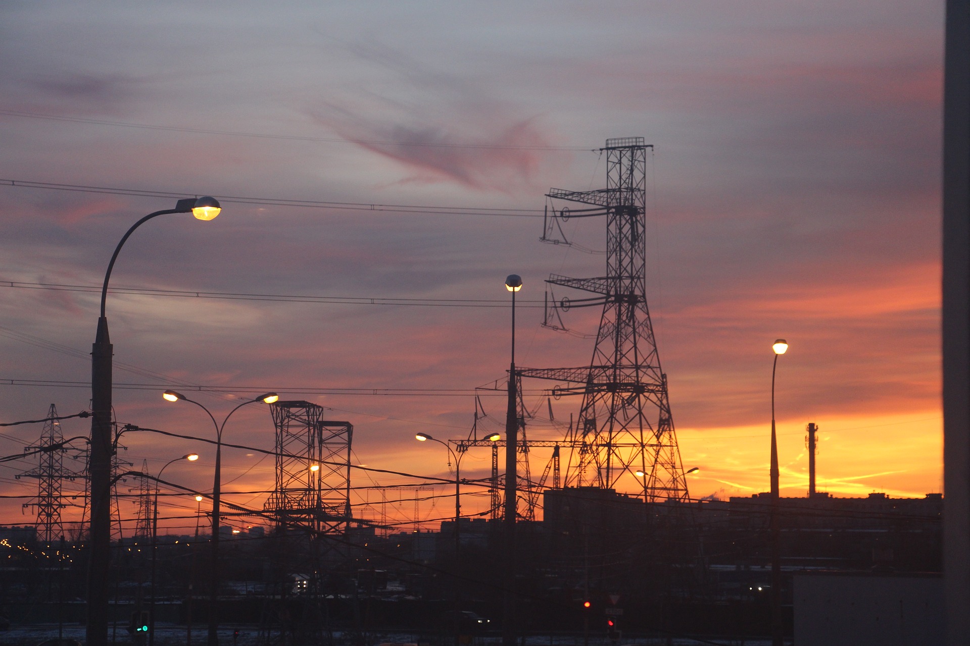 Na imagem: Pôr do sol ao fundo de torres com linhas de transmissão de energia, com alguns postes com luzes acesas (Foto: Inna/Pixabay)