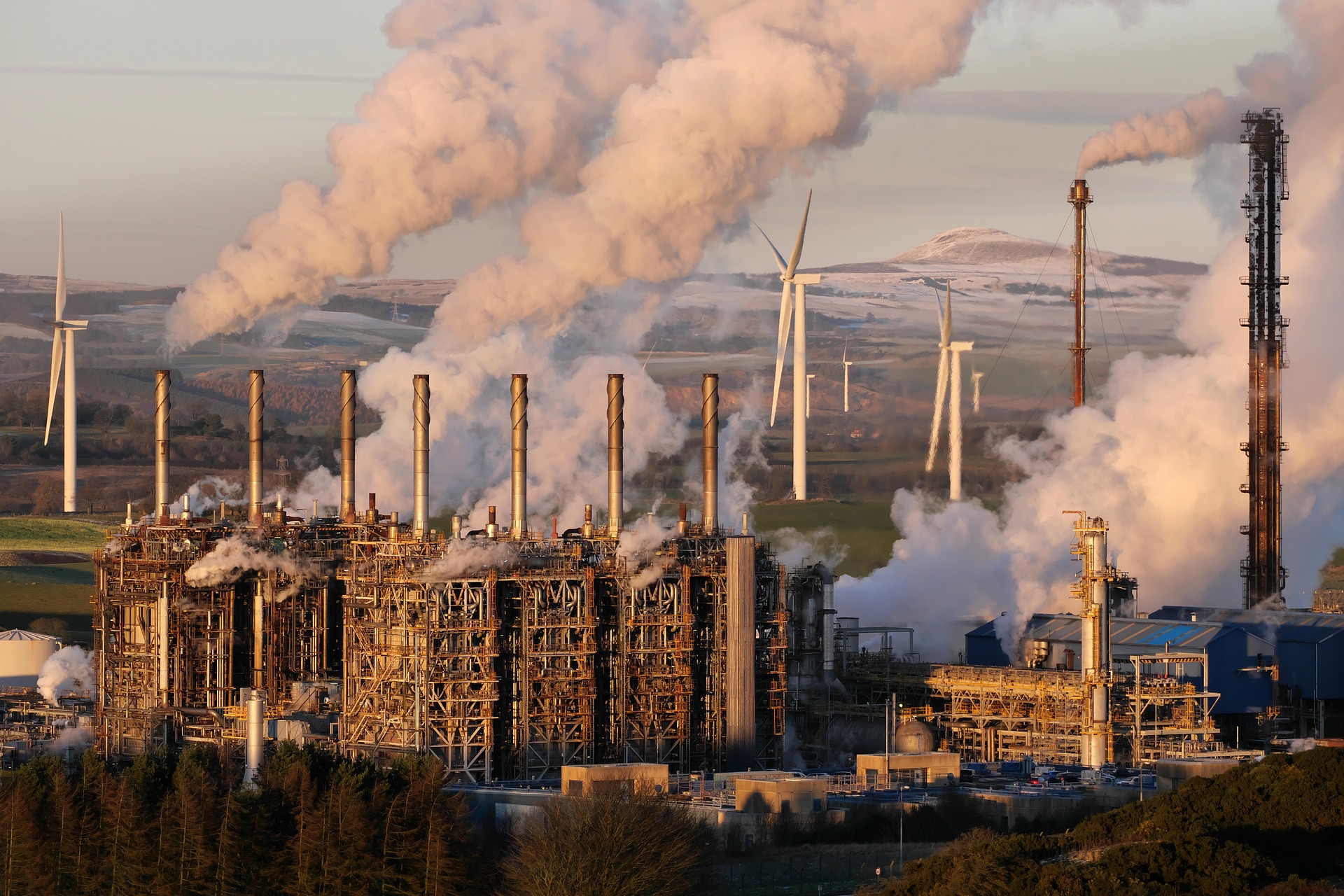 Combustíveis fósseis lideram casos de greenwashing no entre bancos e empresas do setor financeiro. Na imagem: Chaminés de indústria de petróleo emitindo grande volume de gases de efeito estufa, gerando uma densa fumaça branca e, ao fundo, turbinas eólicas (Foto: Emphyrio/Pixabay)