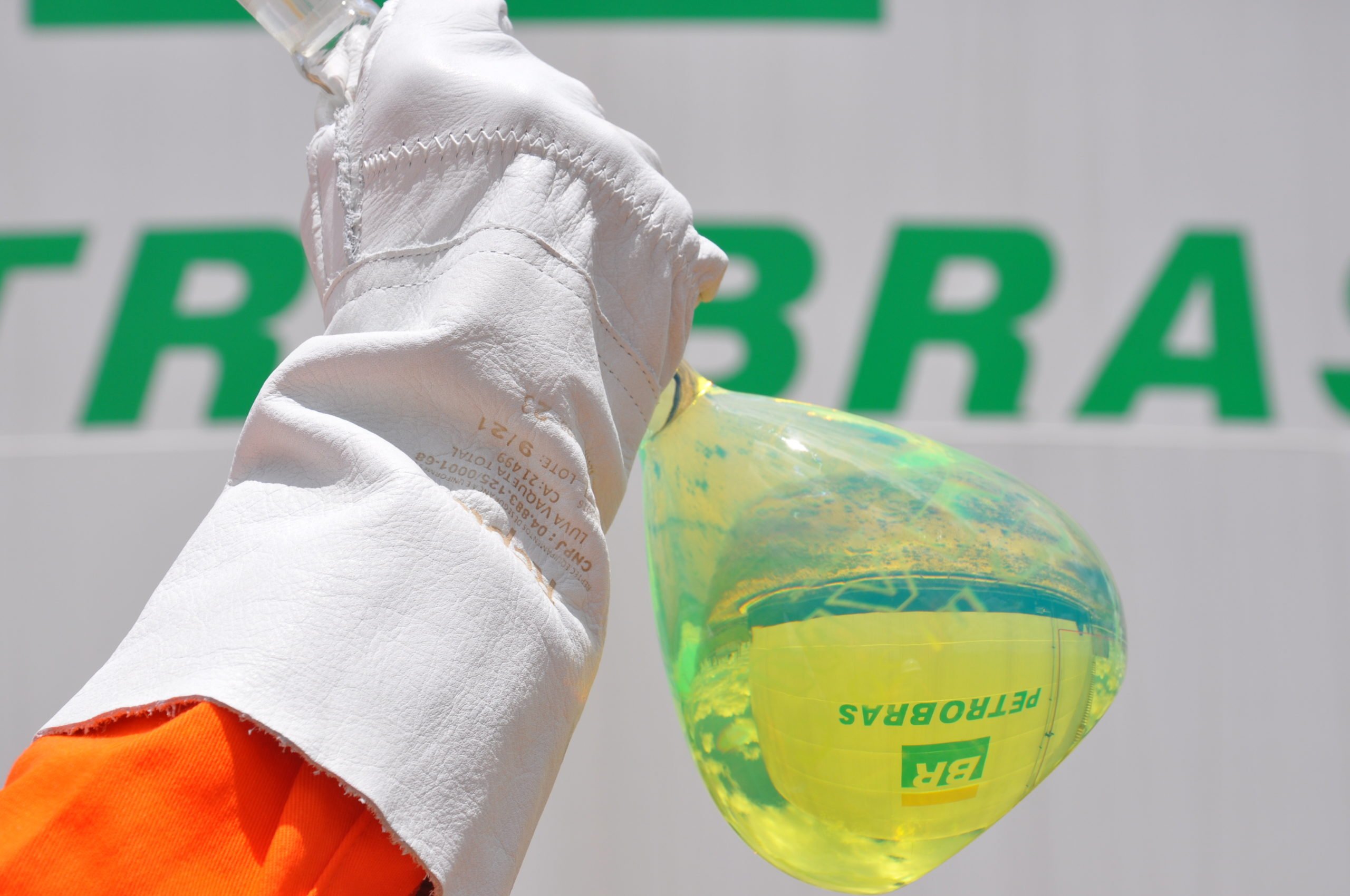 Na imagem: Mão, com luva de proteção branca, segura frasco de laboratório, de vidro, contendo amostra de diesel com teor renovável, da Petrobras, um líquido esverdeado e translúcido; ao fundo, tanque de armazenamento de combustíveis (Foto: Michel Chedid/Petrobras)