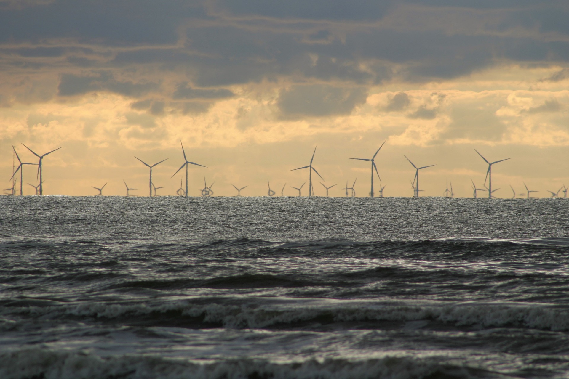 Na imagem: Ondas na praia, em primeiro plano, e, ao fundo, muitas turbinas eólicas em fazenda eólica offshore (Foto: David Will/Pixabay)
